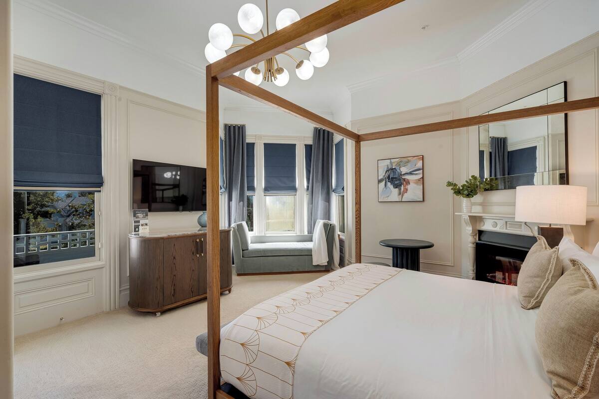 A hotel room with a four-poster bed, a television, a seating area with a sofa and curtains, and a large mirror.