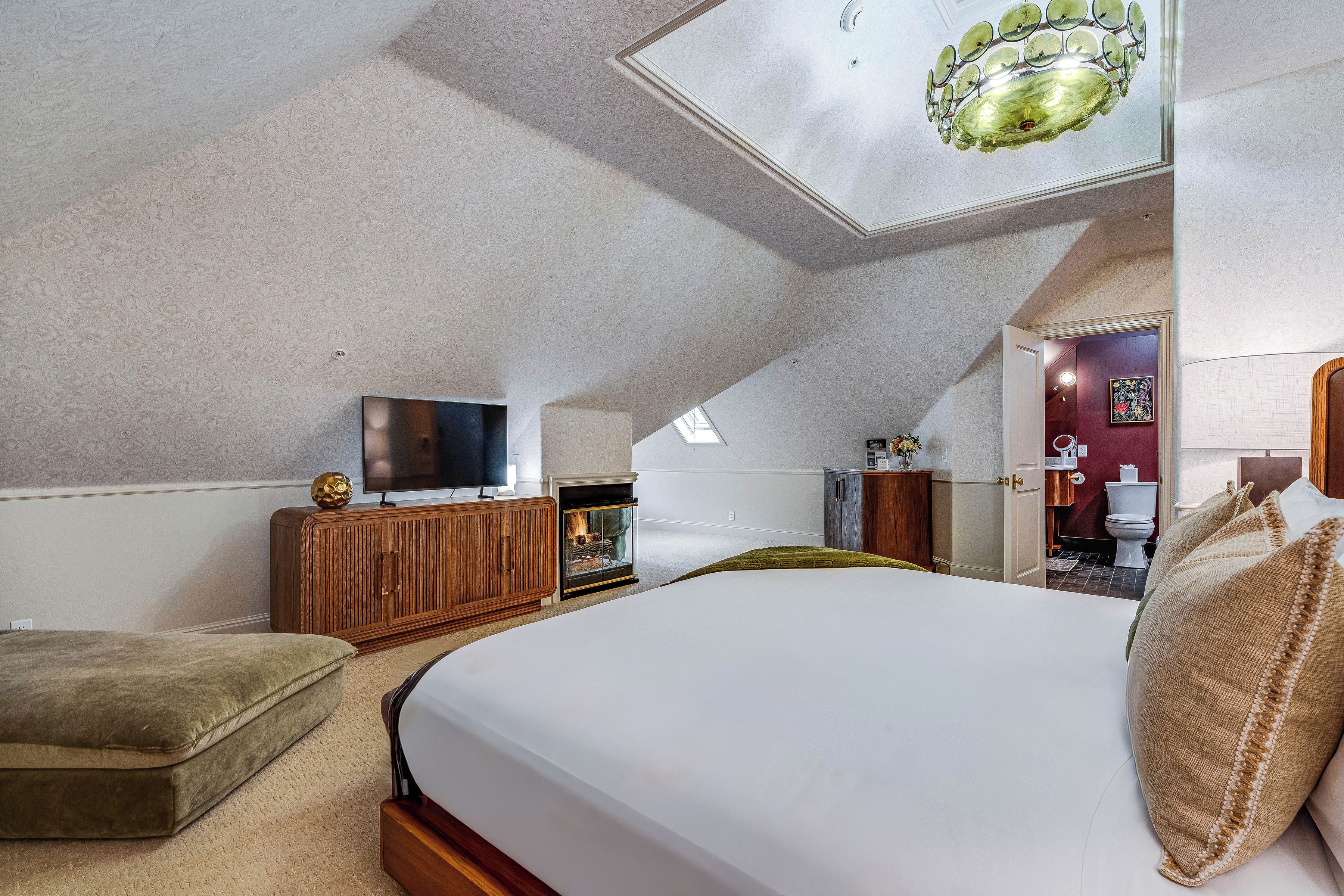 A bedroom with a large bed, a television, a fireplace, and a view of the bathroom.