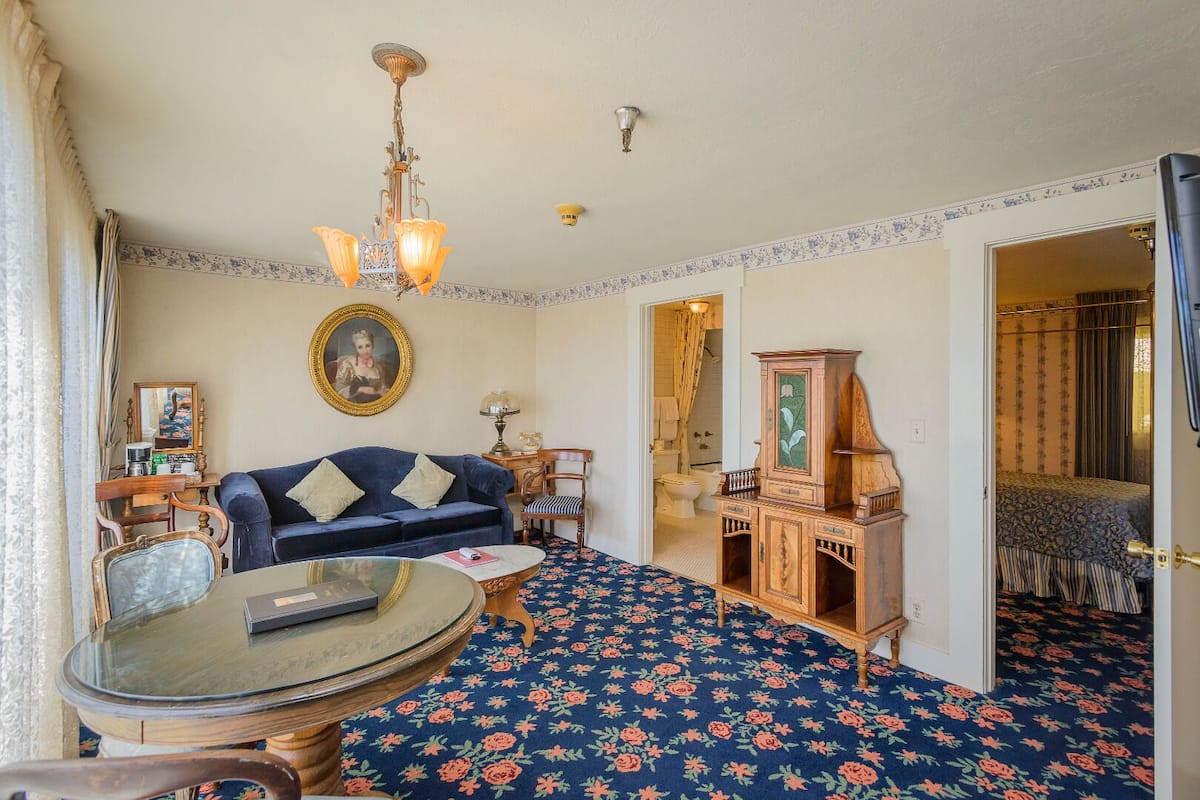 Ein Zimmer mit blauem floralem Teppich, einem dunkelblauen Sofa, einem runden Glastisch, einem Holzschrank und einer Kristallleuchtre.