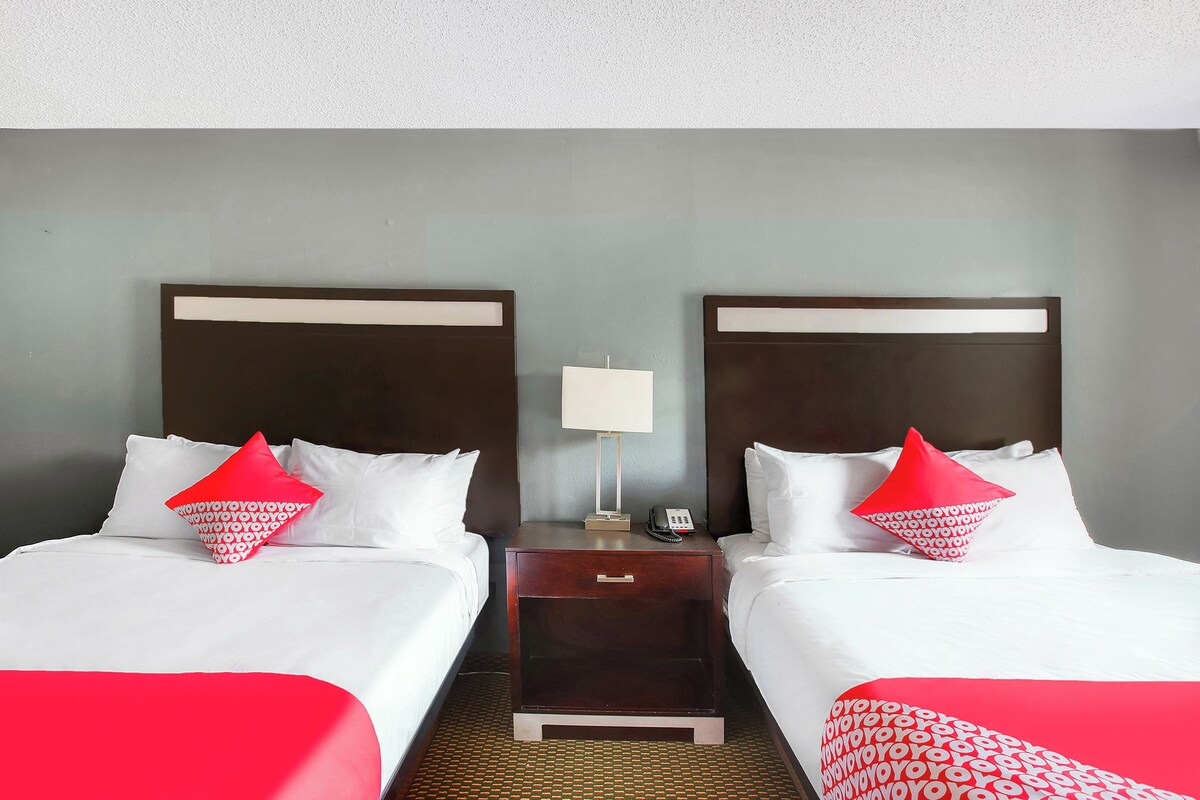 Habitación de hotel con dos camas, cada una con ropa de cama blanca y almohadas rojas, una mesita de noche con lámpara y teléfono, y una alfombra estampada.