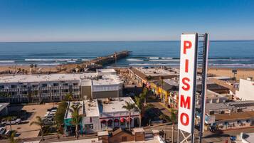 A coastal town with a prominent pier extending into the ocean, a large billboard with the word "PISMO" on a tall structure, and a mix of commercial and residential buildings along the beachfront.