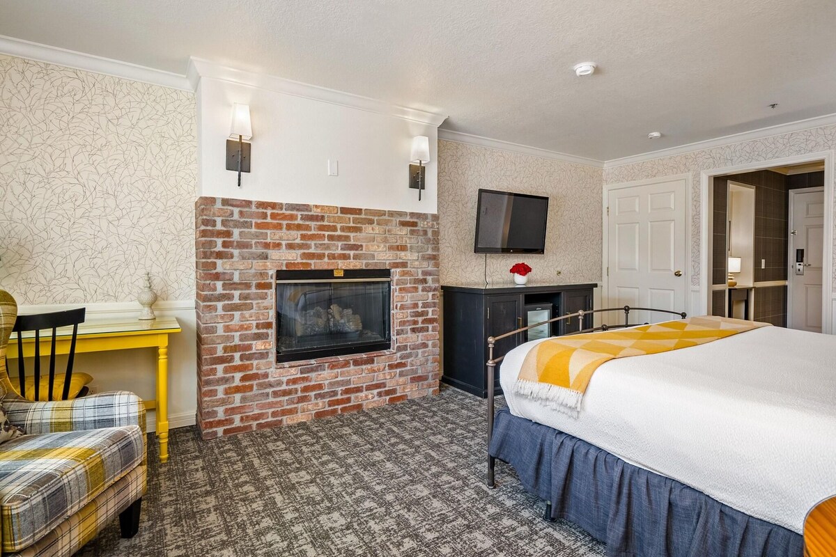 Ein Hotelzimmer mit Kamin aus Backsteinen, einem Bett mit gelb-weißer Decke, einem Schreibtisch mit Stuhl und einem an der Wand befestigten Fernseher.