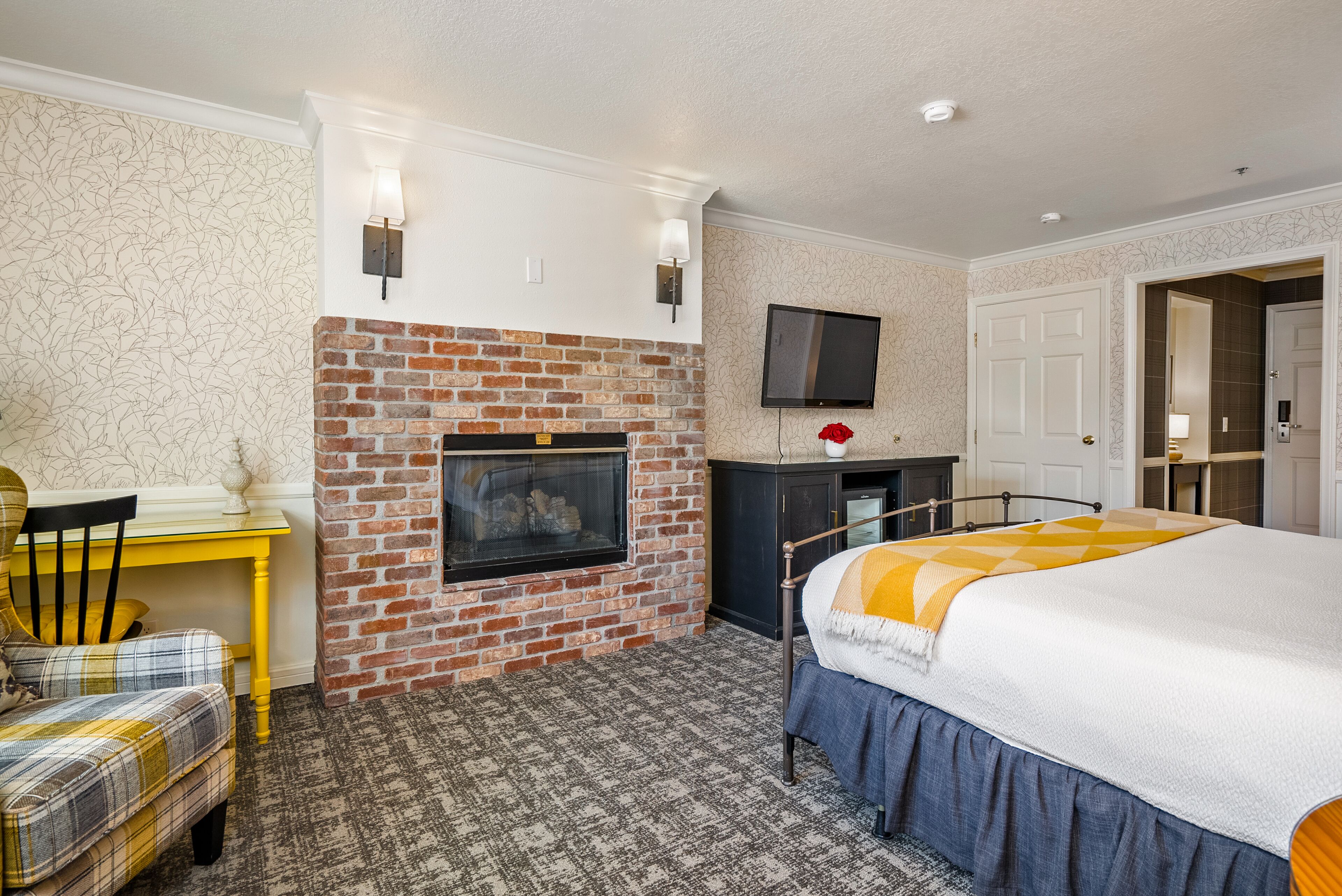 A hotel room with a brick fireplace, a flat-screen TV mounted above it, a bed with a blue skirt, and a yellow table with a plaid chair.