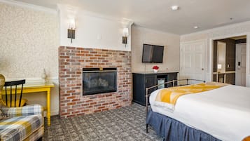 A hotel room with a brick fireplace, a flat-screen TV mounted above it, a bed with a blue skirt, and a yellow table with a plaid chair.