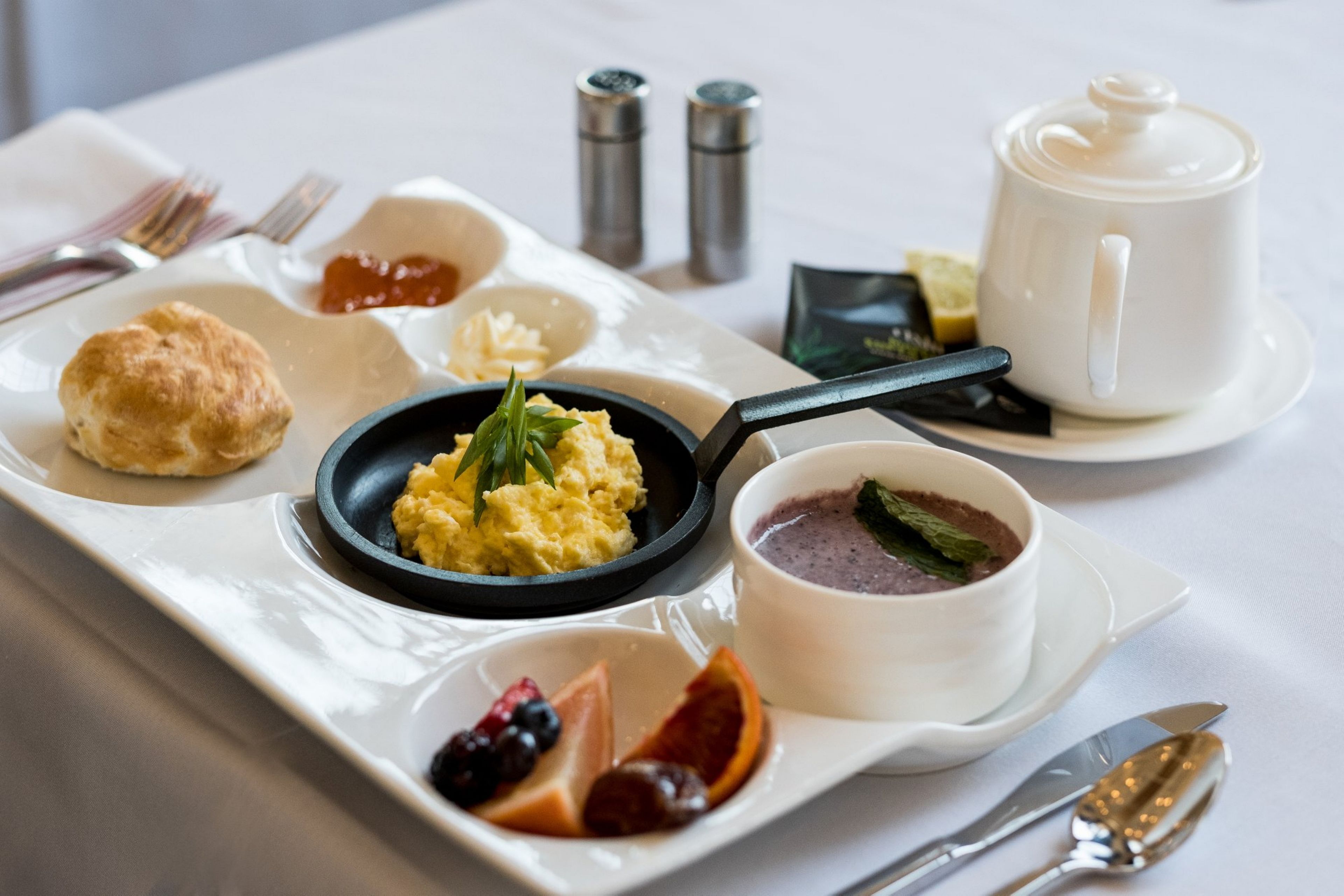 A breakfast tray with scrambled eggs, a bowl of fruit compote, a biscuit, a cup of coffee, and a small bowl of dessert.