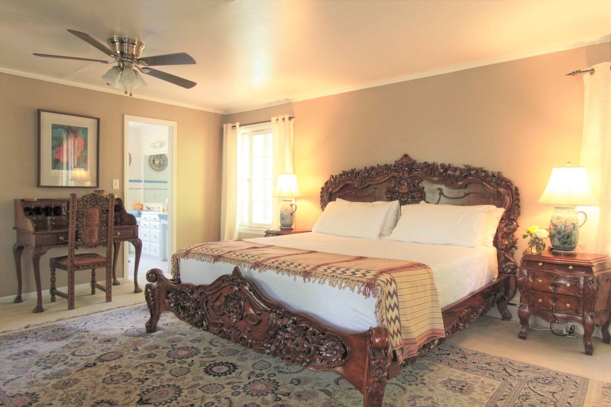 A bedroom with a large ornate bed, a ceiling fan, a wooden nightstand with a lamp, and a chair.