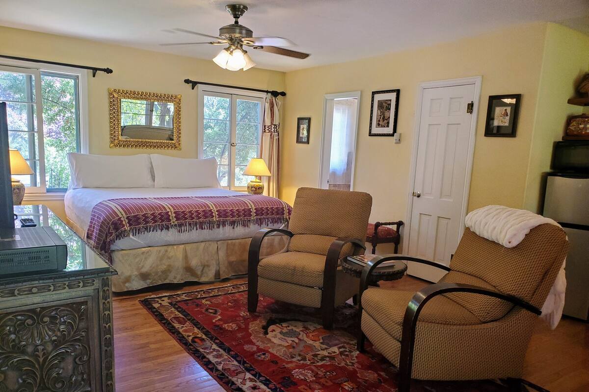 A bedroom with a bed, two chairs, a desk, a ceiling fan, and a door.