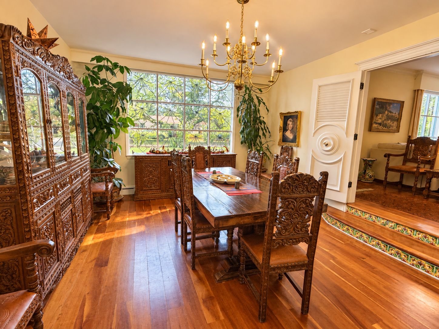 A dining room with a wooden table, chairs, a chandelier, and a large window with a view of greenery.