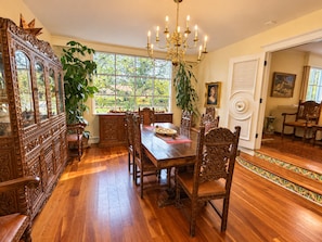 A dining room with a wooden table, chairs, a chandelier, and a large window with a view of greenery.