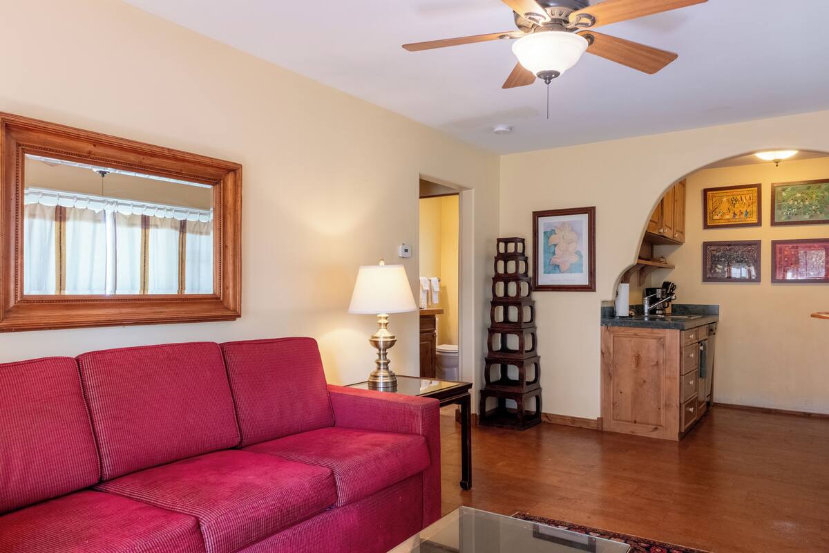 A living room with a red sofa, a ceiling fan, a lamp, and a kitchen area with wooden cabinets and a sink.