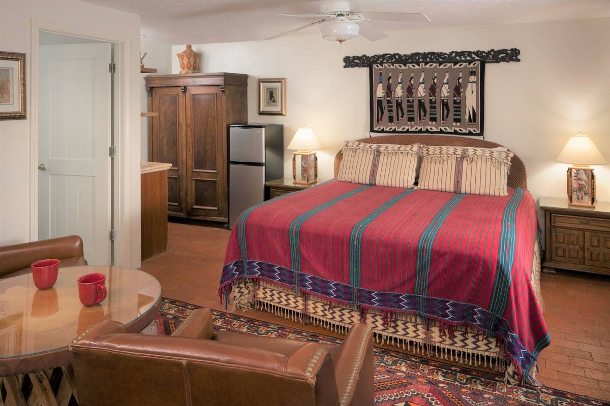 A hotel room with a bed, a refrigerator, a dining table with chairs, and a ceiling fan.