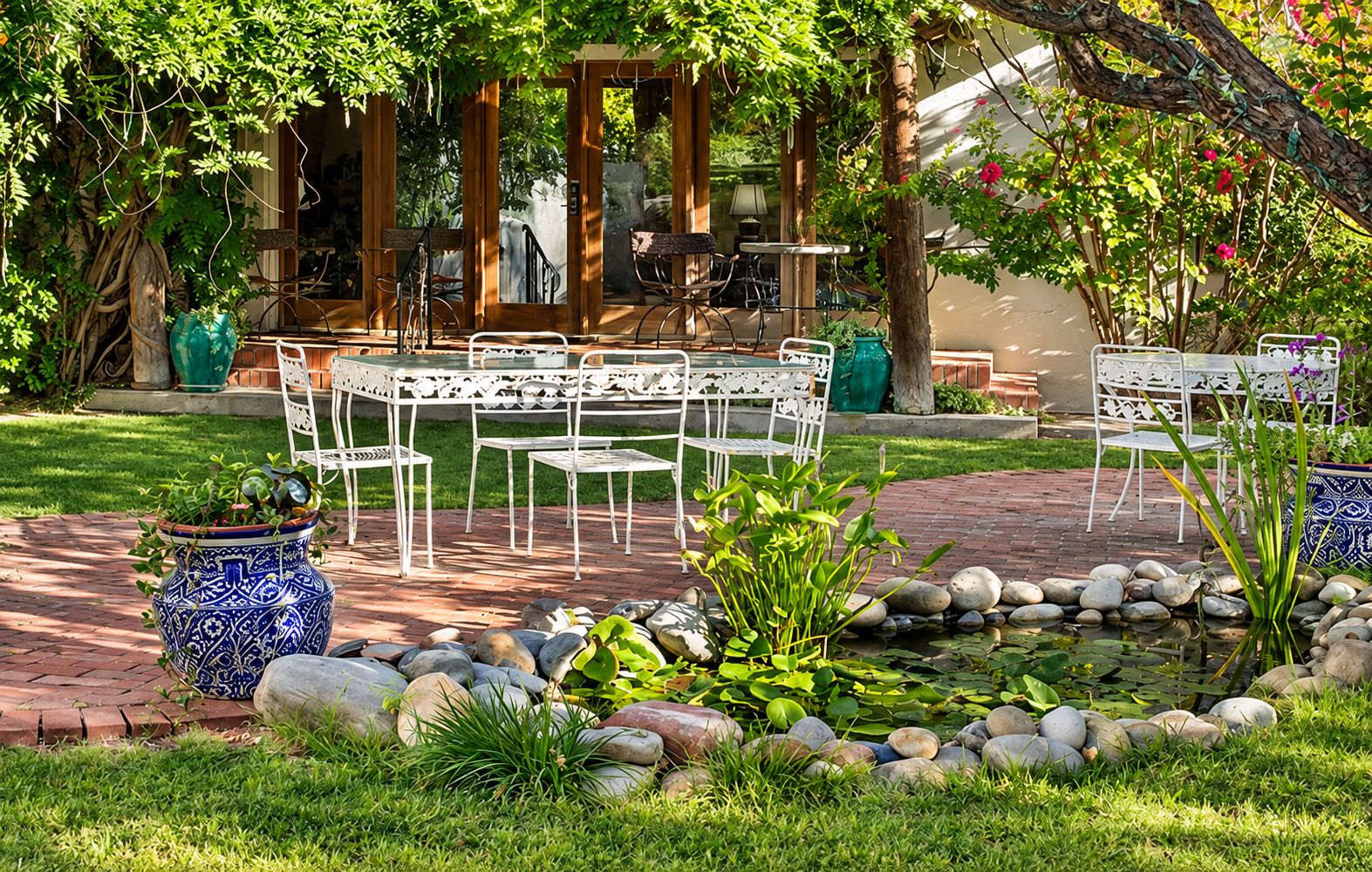 An outdoor seating area with a brick pathway, white metal chairs, and a small water feature with rocks.
