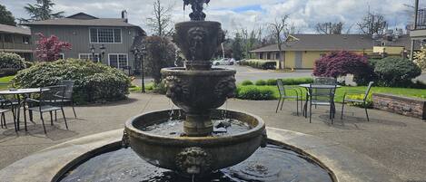 A stone fountain with water spouting from its top, surrounded by a circular basin. In the background, there are residential buildings, a street, and a small outdoor seating area with chairs and tables.