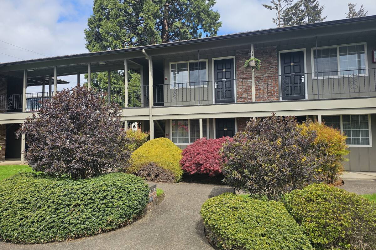 A two-story building with a covered walkway and a garden area with various shrubs.