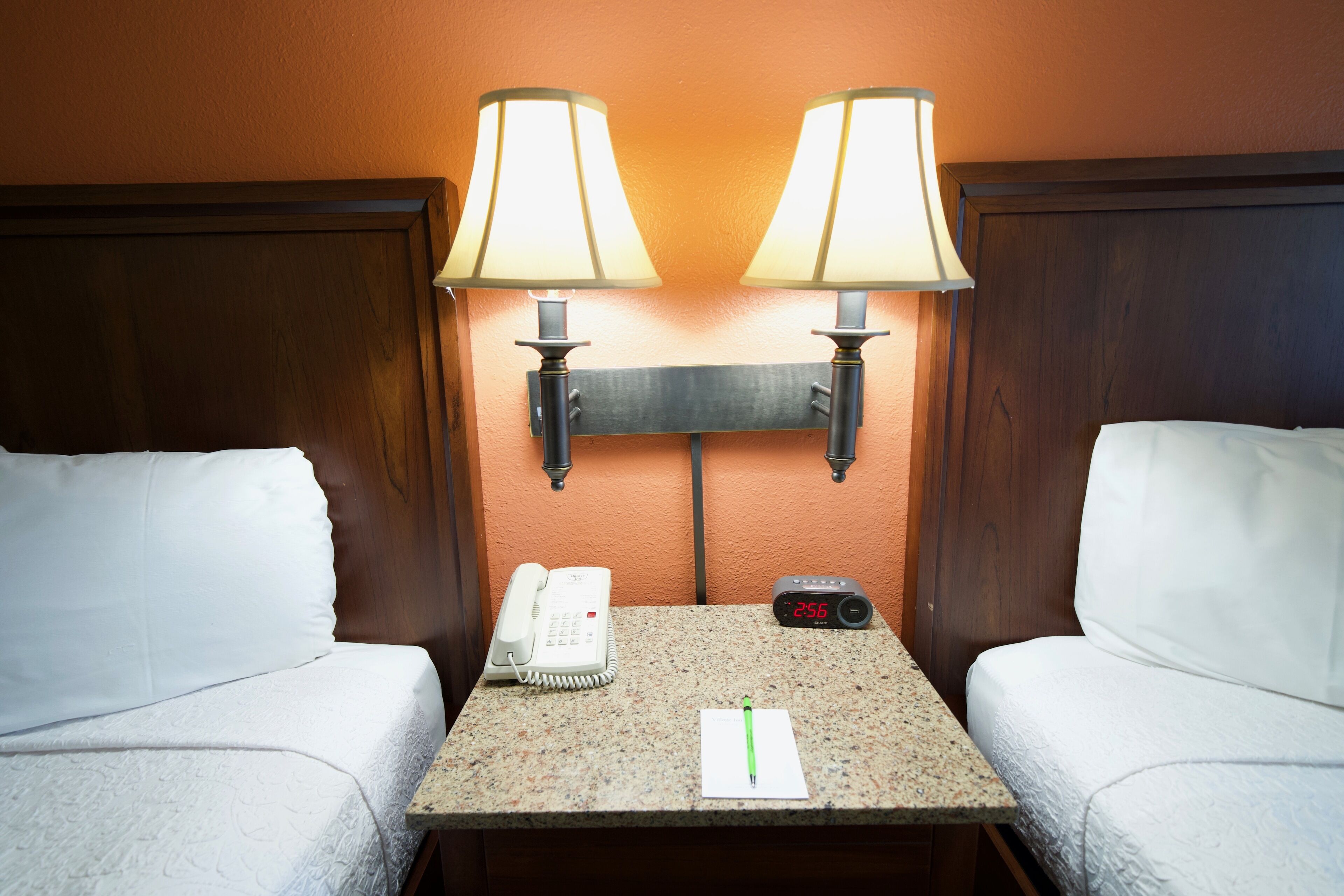 A hotel room with two beds, a nightstand with a phone, a digital clock, and two wall-mounted lamps.