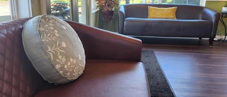 A living room with a brown leather sofa, a grey cushion, and a yellow pillow. There is a window with a view of buildings and trees.