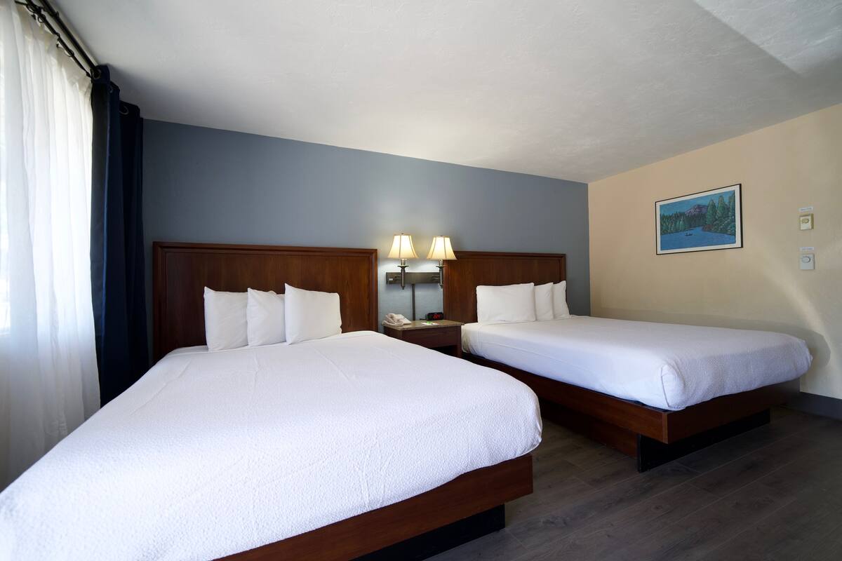 A hotel room with two single beds, wooden headboards, a nightstand with lamps, a painting on the wall, and a window with curtains.