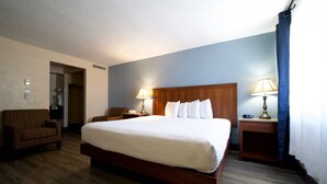 Una habitación de hotel con una cama grande, dos lámparas de noche, una mesita de noche de madera, un sillón a rayas y una puerta que da al baño.