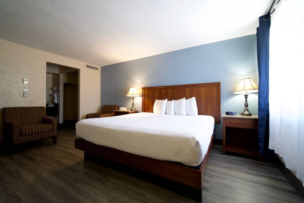 A hotel room with a large bed, two bedside lamps, a wooden nightstand, a striped armchair, and a door leading to a bathroom.