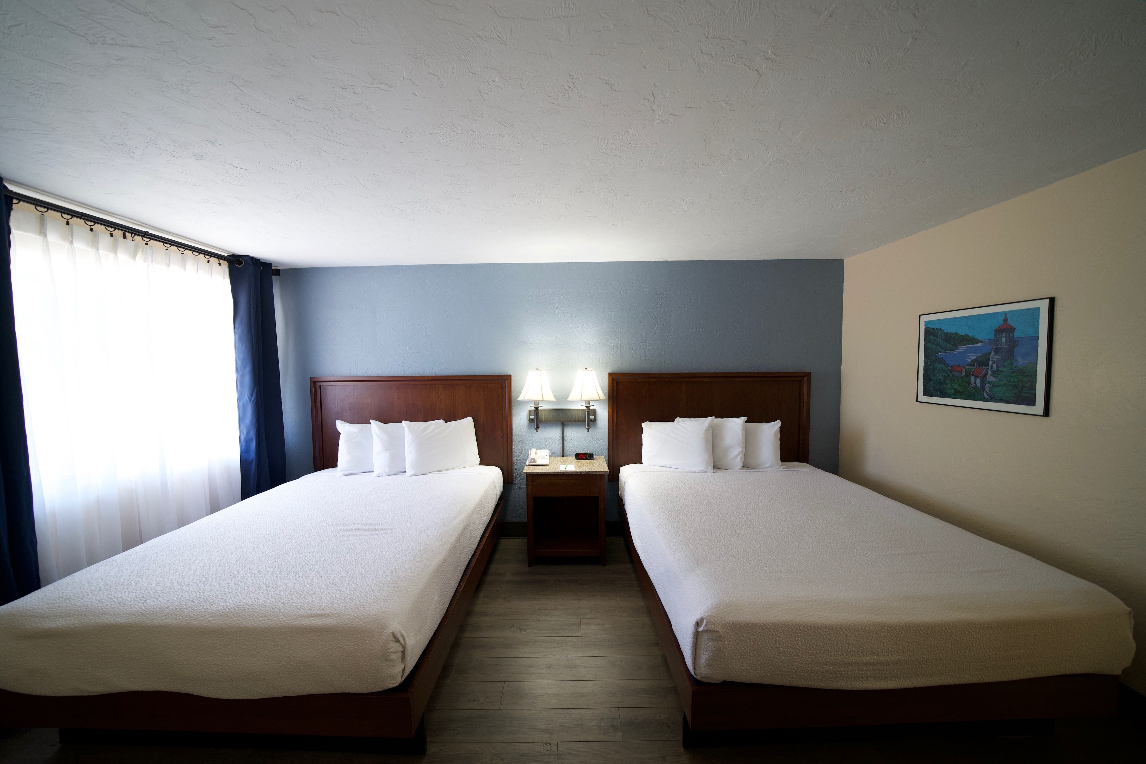 Une chambre d’hôtel avec deux lits simples, une table de chevet avec des lampes, un tableau au mur et une fenêtre avec des rideaux.