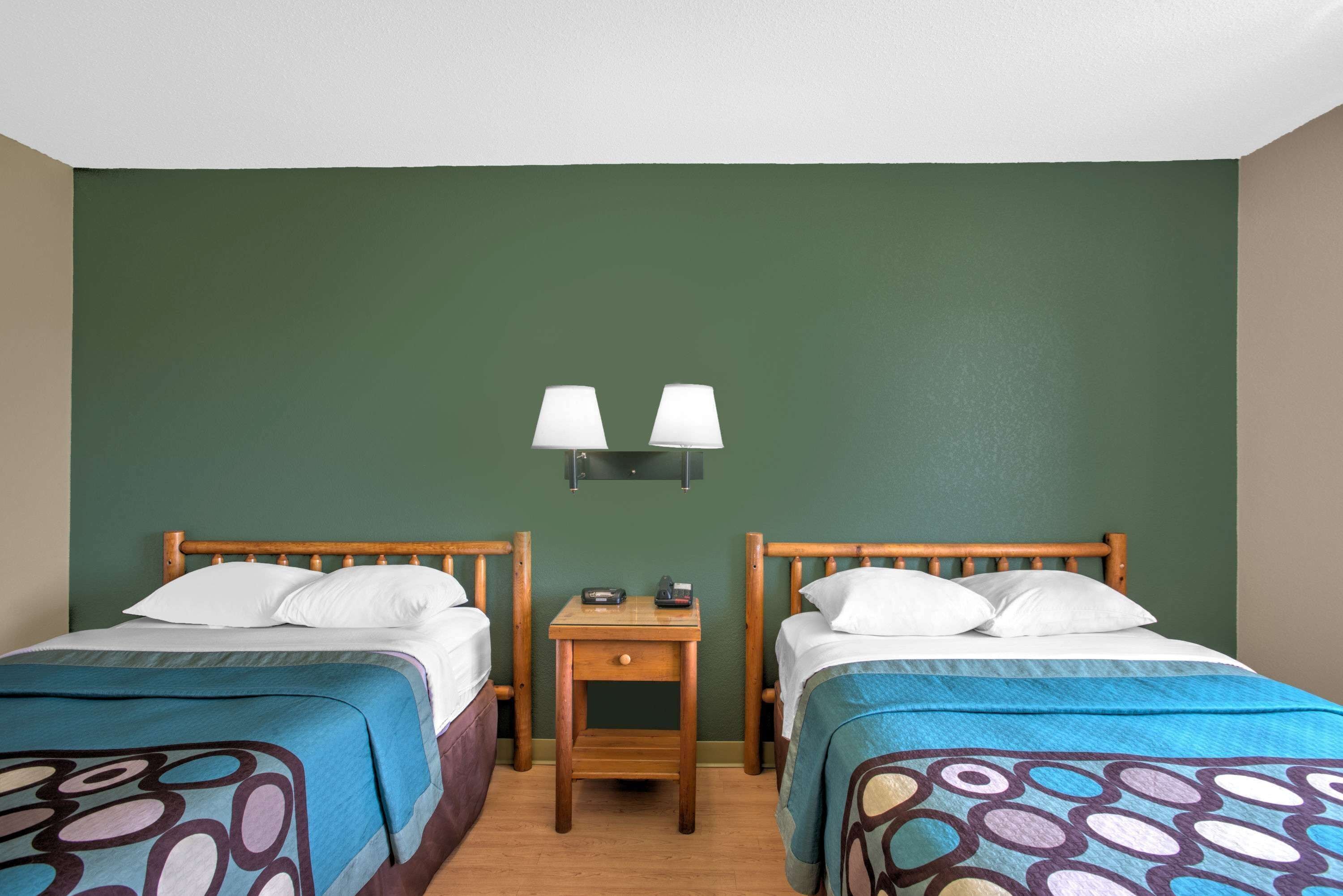 A room with two beds, each with a wooden headboard, a bedside table with a lamp, and a green wall.