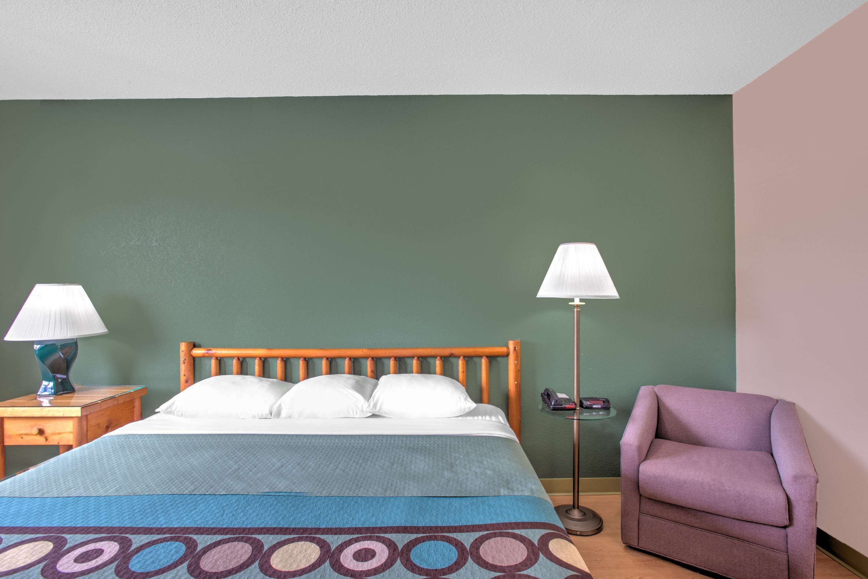 Una cama bien hecha con colcha azul y marrón estampada, dos lámparas de noche, una mesita de noche de madera, un sillón morado y una pared verde.