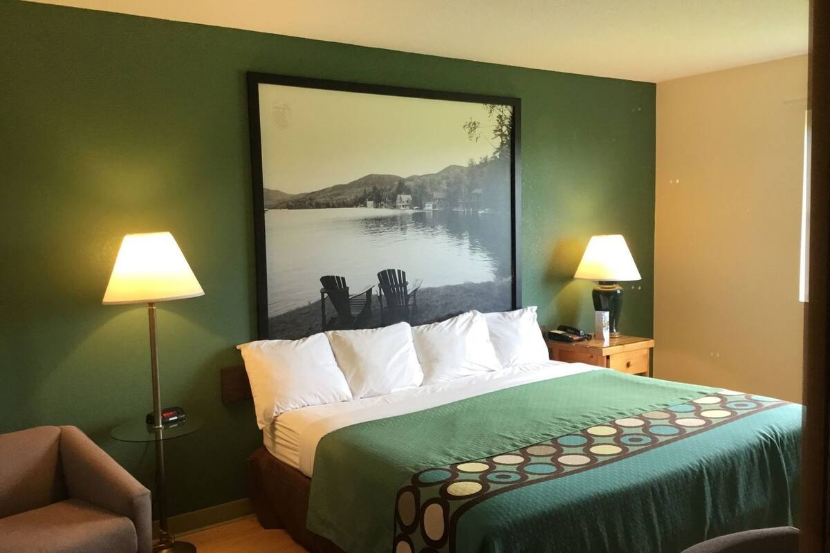 A hotel room with a bed, two lamps, a chair, and a large framed picture of a lake.