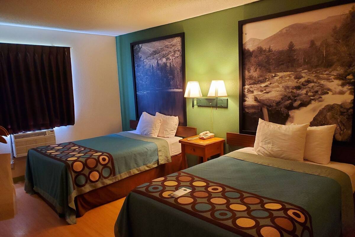 A hotel room with two beds, a nightstand with a phone, and a large framed picture of a mountainous landscape.