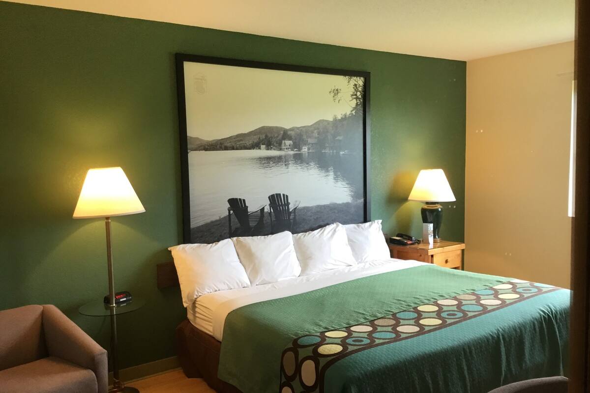 A hotel room with a bed, two lamps, a chair, and a large framed picture of a lake.
