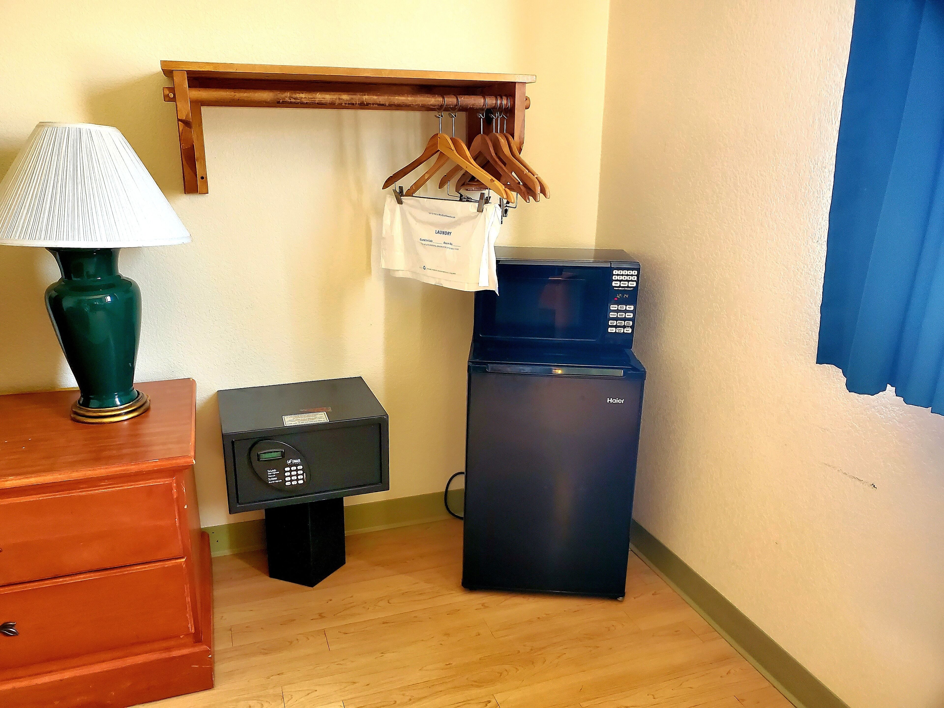 A room with a wooden dresser, a green lamp, a black refrigerator, a microwave, and a wooden shelf with hangers and a white bag.