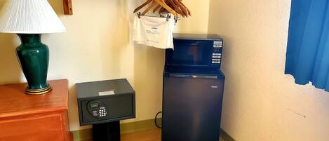 A room with a wooden dresser, a green lamp, a black refrigerator, a microwave, and a wooden shelf with hangers and a white bag.