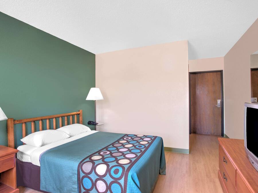 A hotel room with a bed, bedside table, lamp, TV, and a door leading to another room.