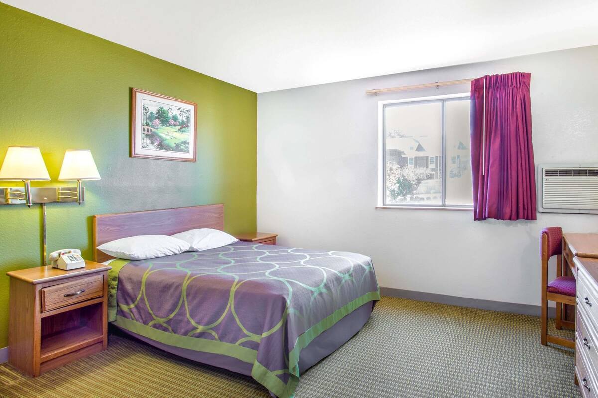 Ein Hotelzimmer mit Bett, Nachttischlampen, einem Nachttisch, einem Sessel, einer Kommode, einem Fenster mit Vorhängen und einem Bild an der Wand.