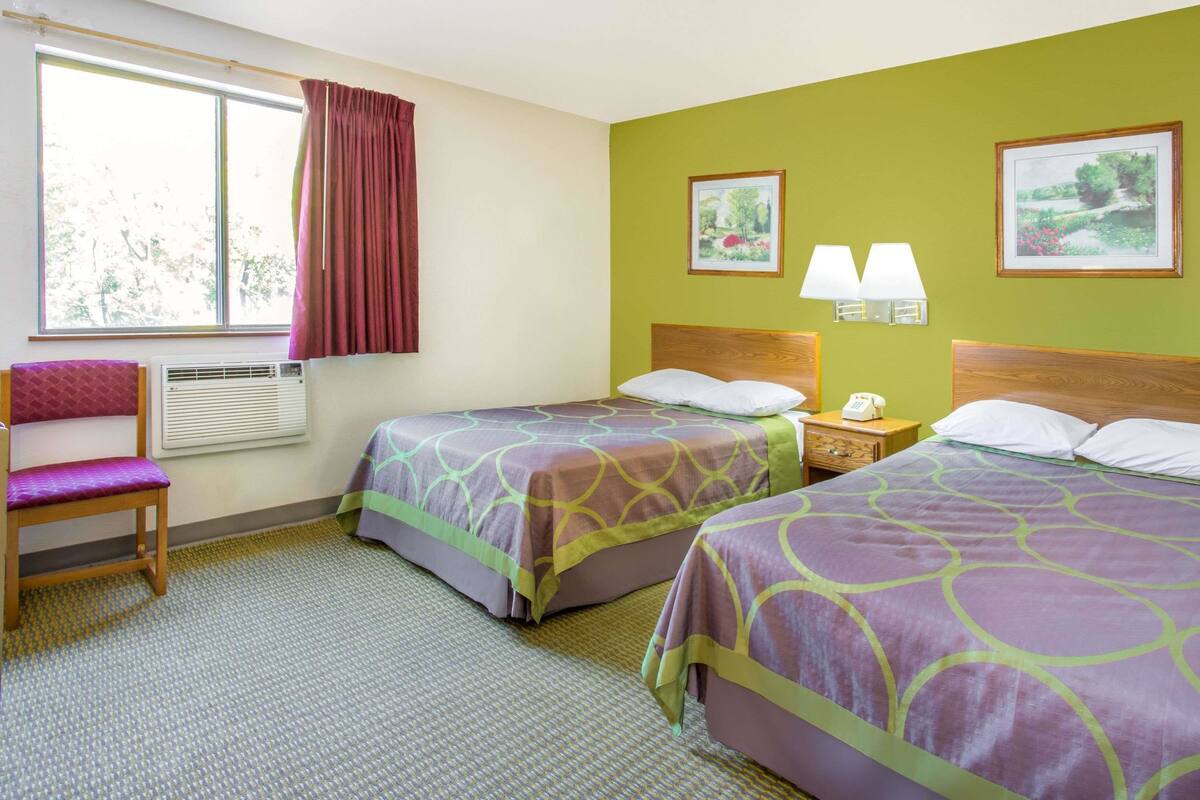 Ein Hotelzimmer mit zwei Betten, einem Sessel, einem Fenster mit Vorhängen und zwei Wandlampen.