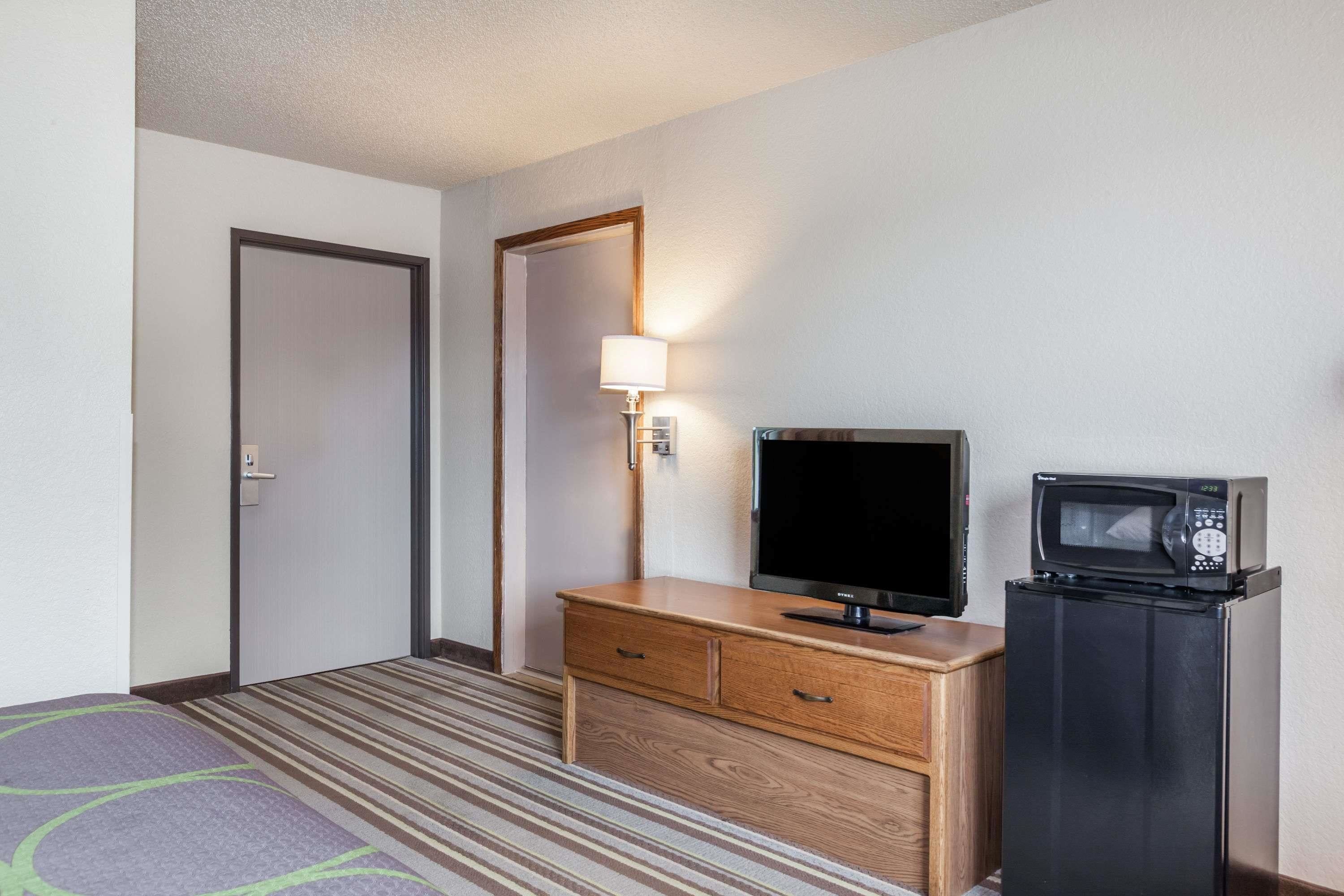 Quarto de hotel com cômoda de madeira, TV de tela plana, micro-ondas, porta e luminária.
