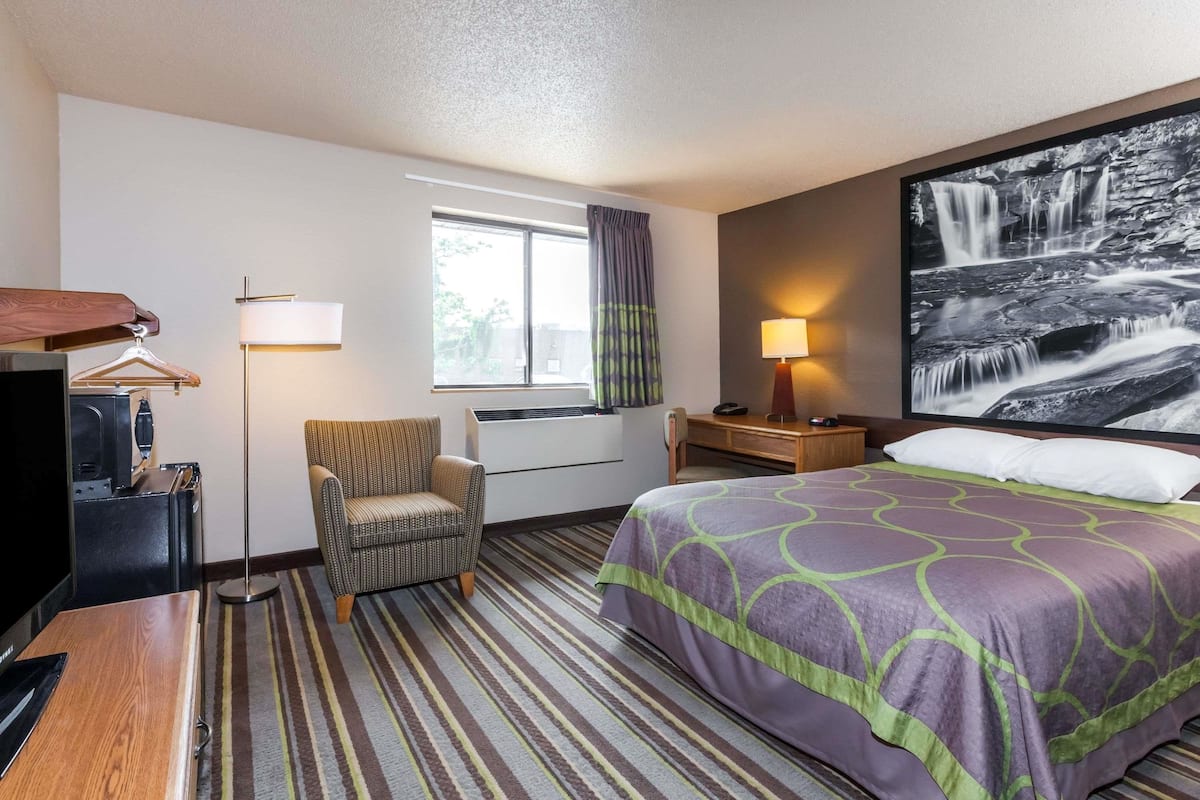 Een hotelkamer met een bed, een stoel, een bureau, een televisie, een lamp en een groot raam met gordijnen.