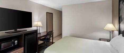 Habitación de hotel con televisión de pantalla plana, escritorio con lámpara, una silla, una cama y mesita de noche con lámpara.