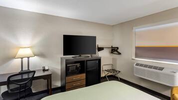 Una habitación de hotel con cama, escritorio con lámpara, televisor, minibar y ventana con persianas.
