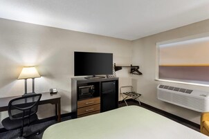 Una habitación de hotel con cama, escritorio con lámpara, televisor, minibar y ventana con persianas.
