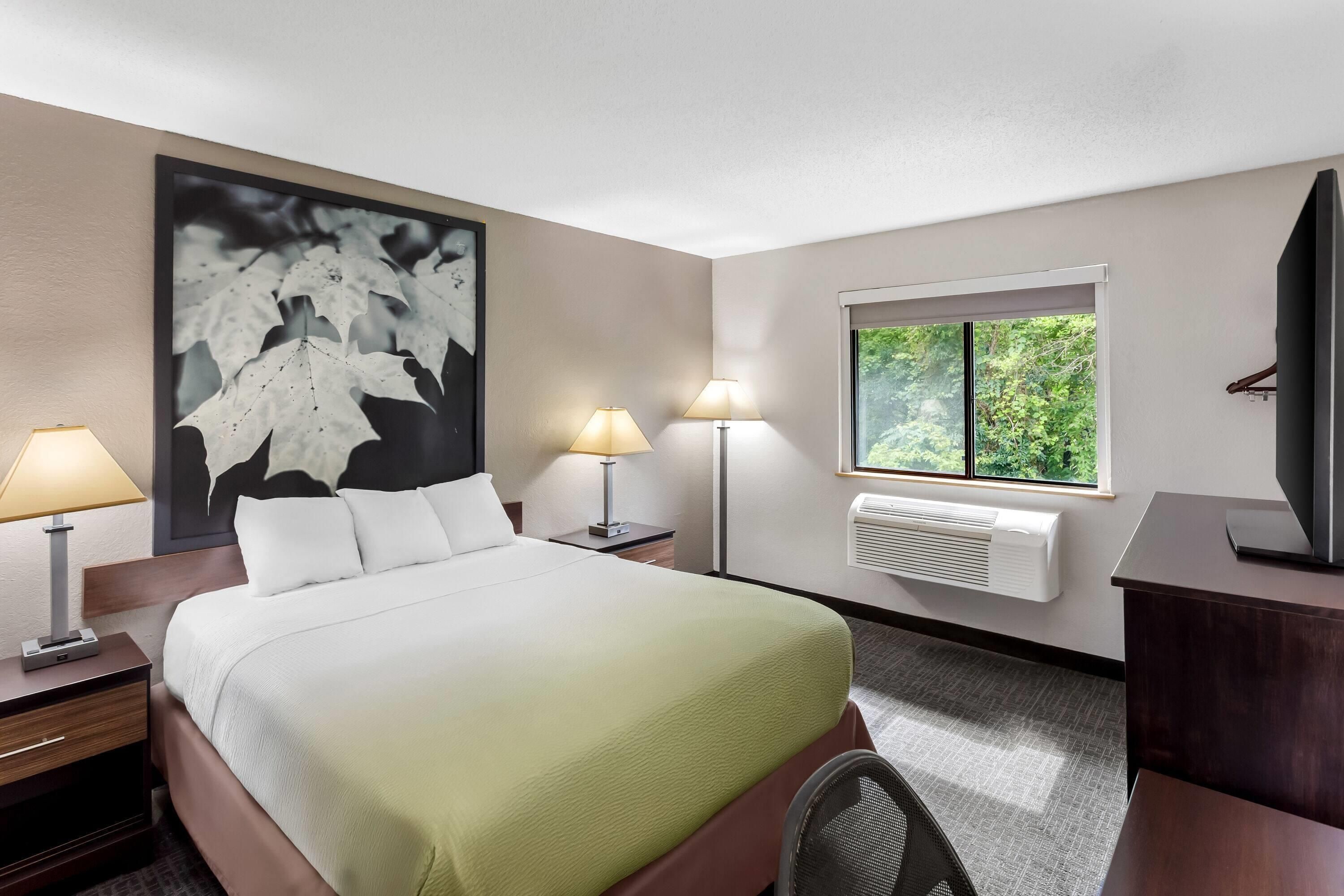 A hotel room with a bed, two bedside lamps, a window with a view, and an air conditioning unit.