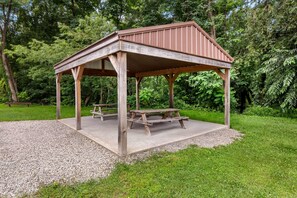 A wooden gazebo with a bench and picnic table, surrounded by gravel and grass, set in a wooded area. - Super 8 by Wyndham Elkins (Elkins)