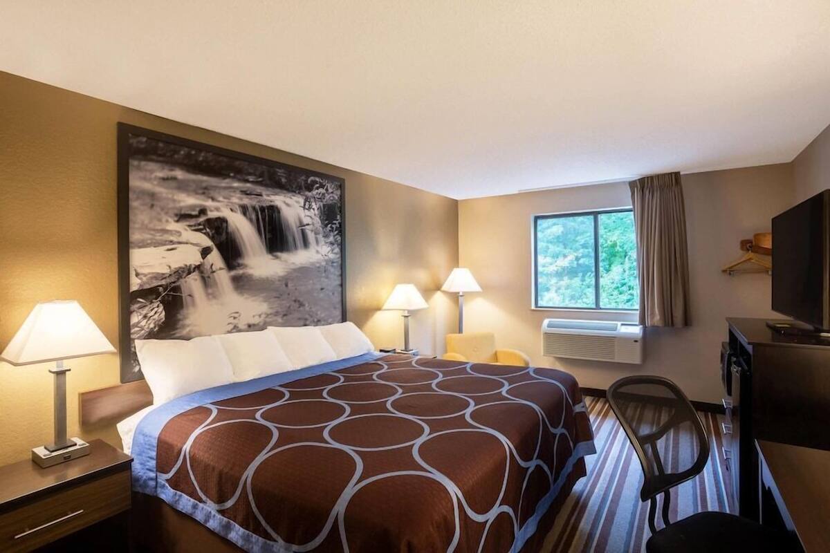 A hotel room with a bed, two bedside lamps, a large window with curtains, a flat-screen TV, and a wall-mounted air conditioning unit.