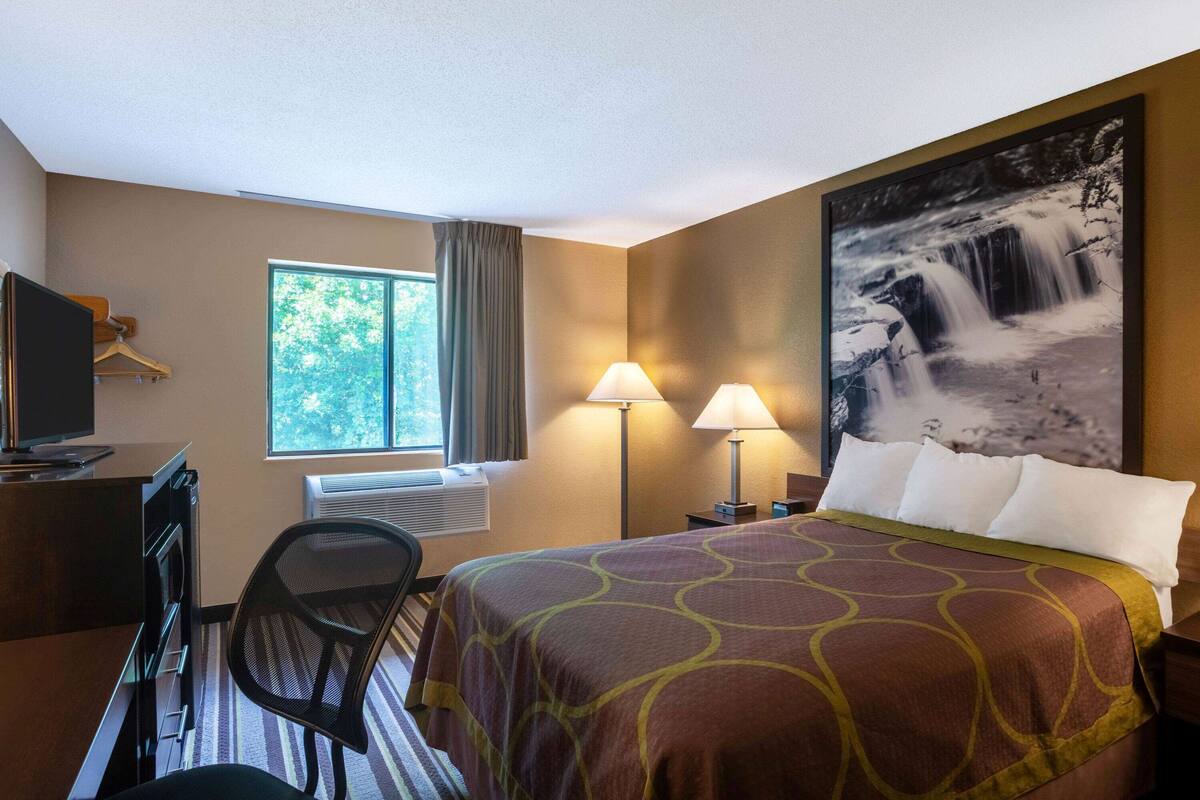 A hotel room with a bed, a desk, a chair, a TV, a window with curtains, two lamps, and a large waterfall painting on the wall.