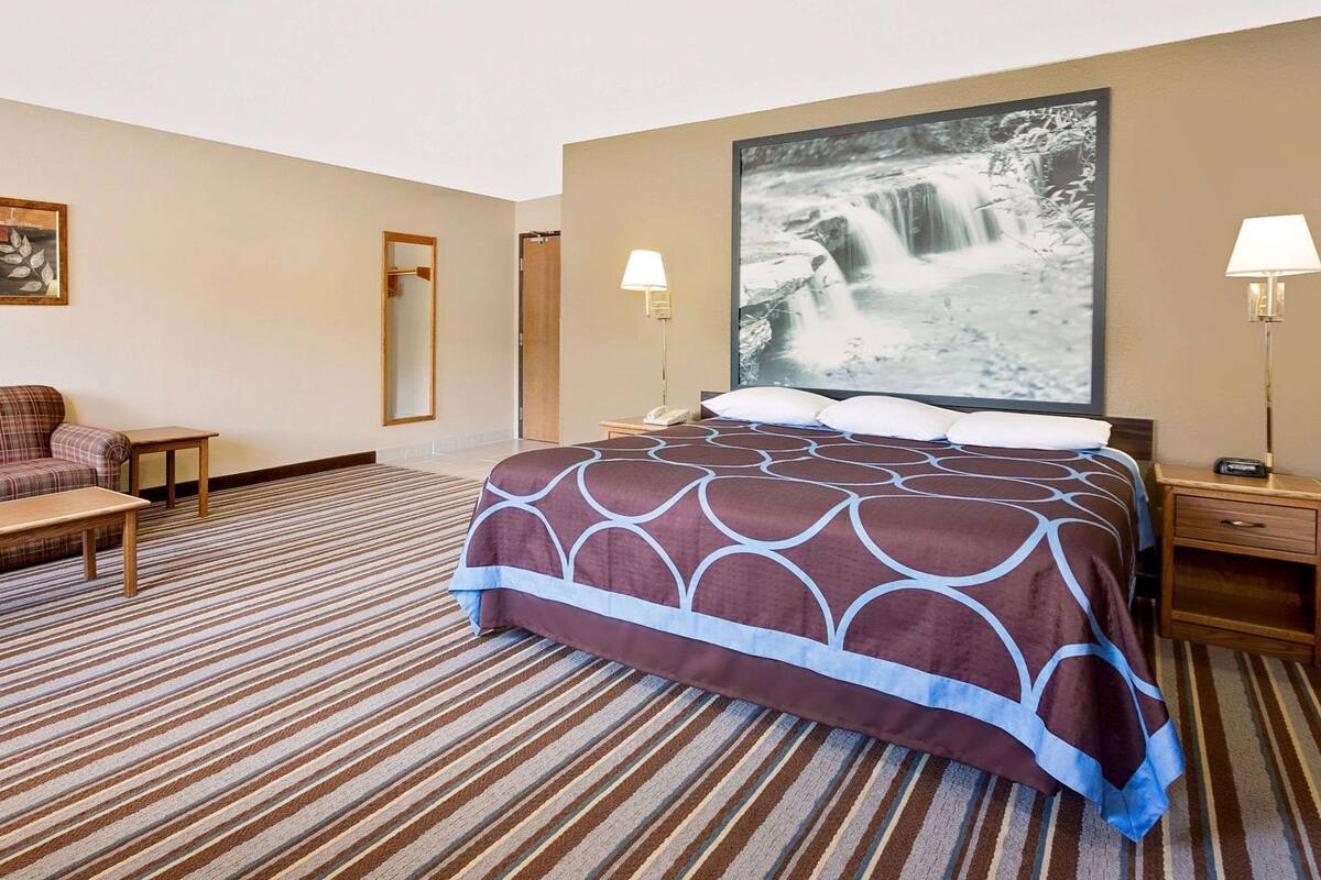 A hotel room with a bed, a sofa, a bench, a nightstand, a lamp, and a large framed picture of a waterfall.