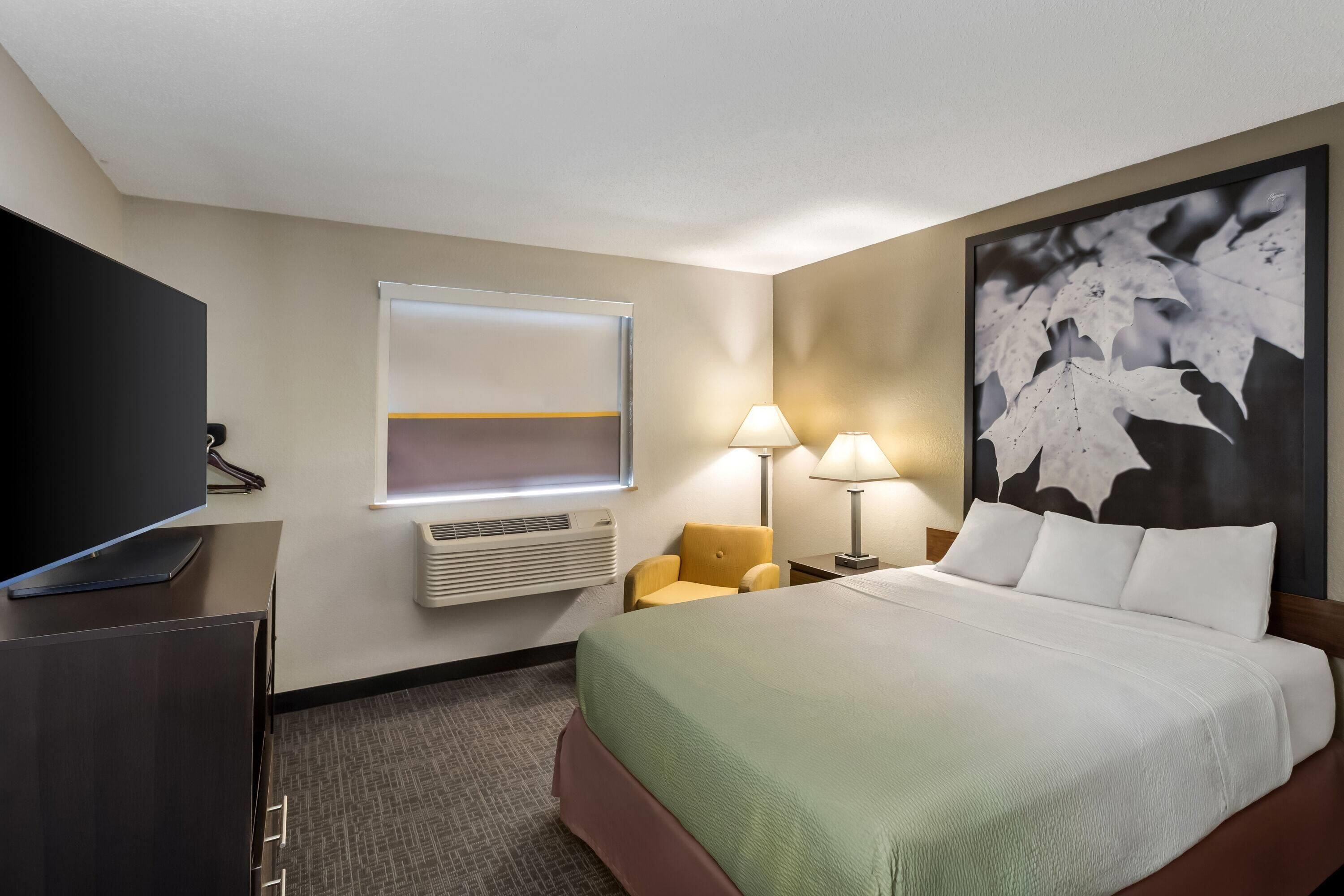 A hotel room with a bed, a television, a window with blinds, a wall-mounted air conditioner, a nightstand with a lamp, and a large abstract painting.