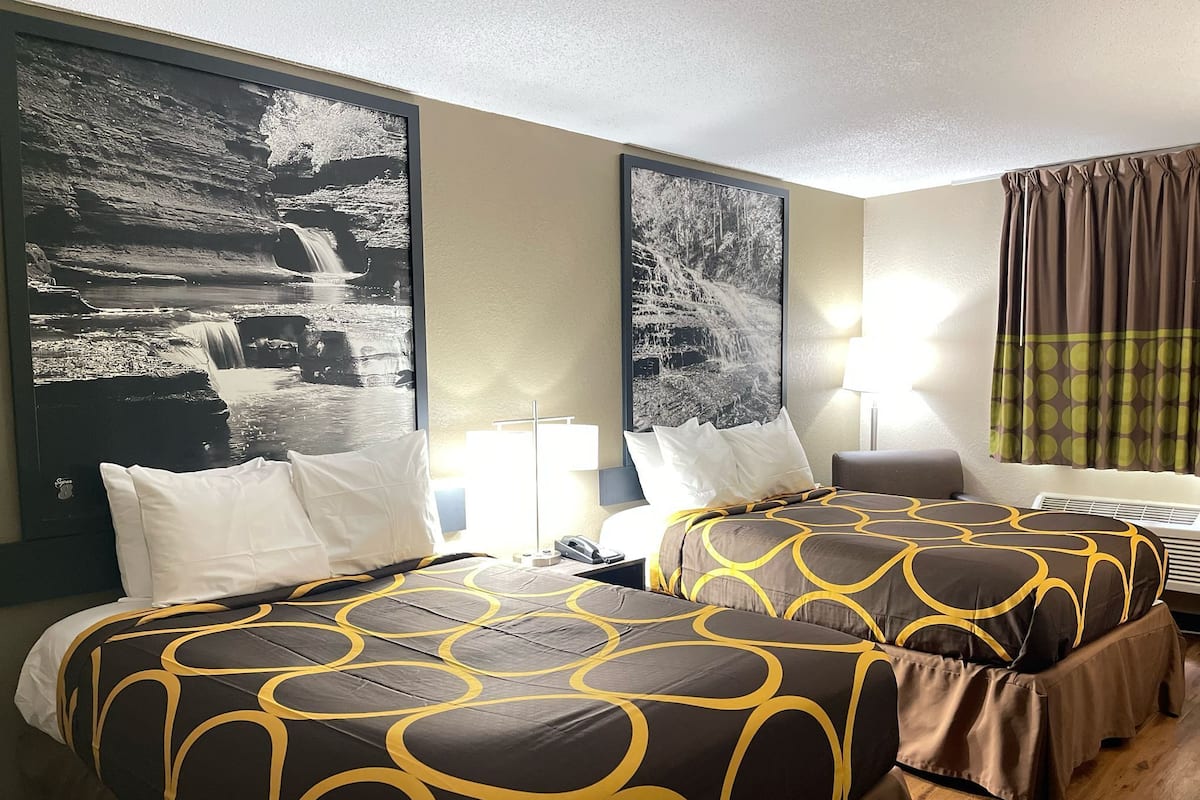 A hotel room with two beds, each with a patterned bedspread, a large wall-mounted picture, a bedside table with a lamp, and a window with curtains.