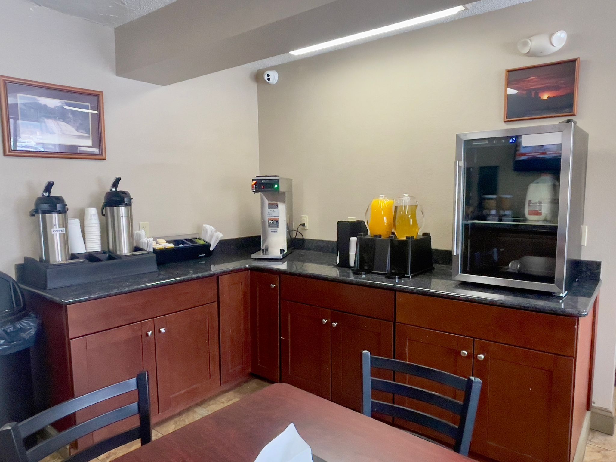 Uma pequena cozinha com máquina de café, geladeira e micro-ondas. Há duas cadeiras e uma mesa no centro.