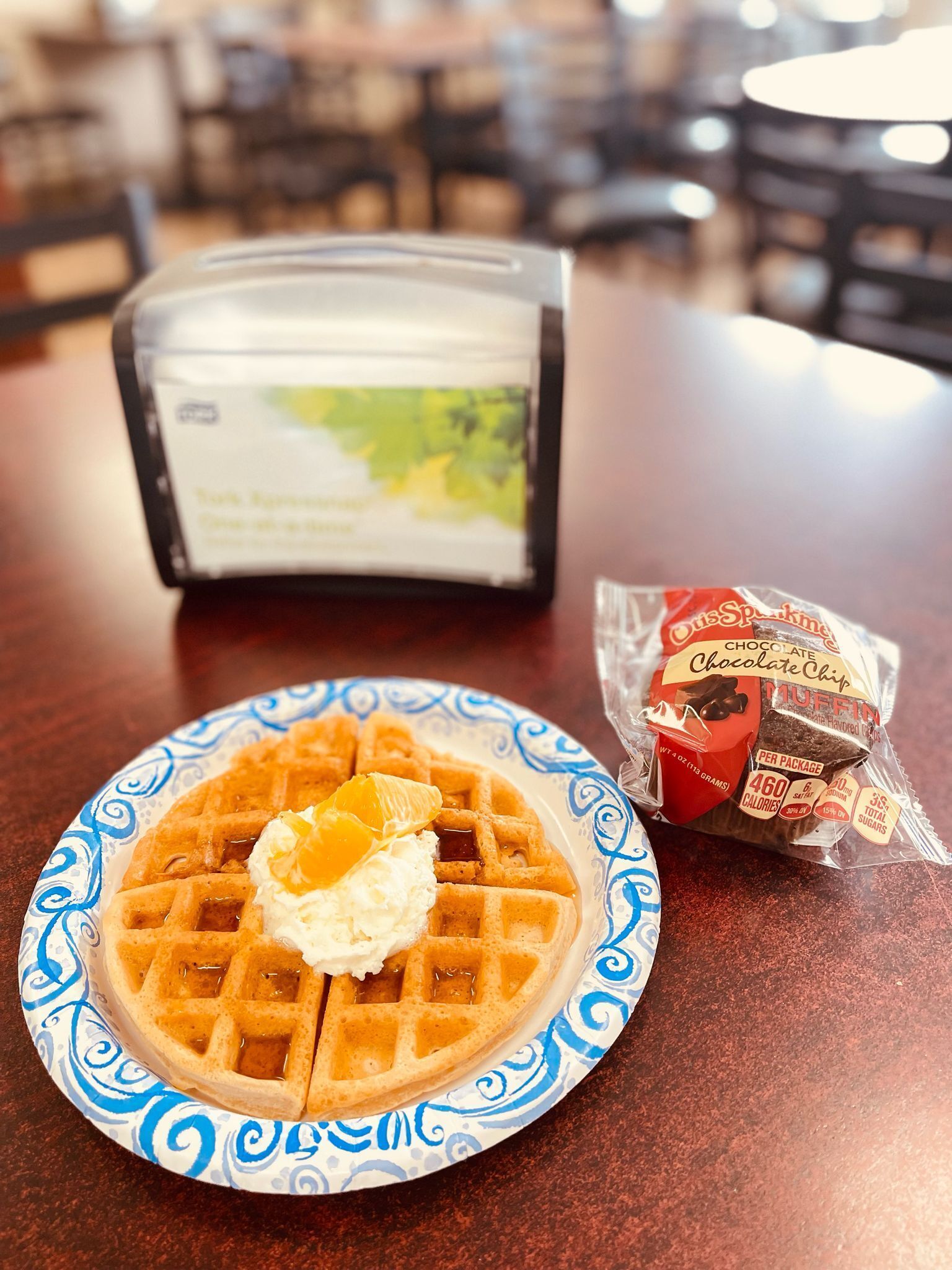 Un plato de waffles con crema batida y jarabe, un muffin de chispas de chocolate y una bolsa de galletas con chispas de chocolate sobre una mesa.