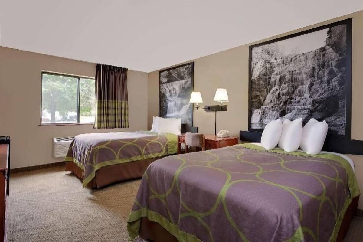 A hotel room with two beds, a large window, a painting of a waterfall, and a nightstand with lamps.