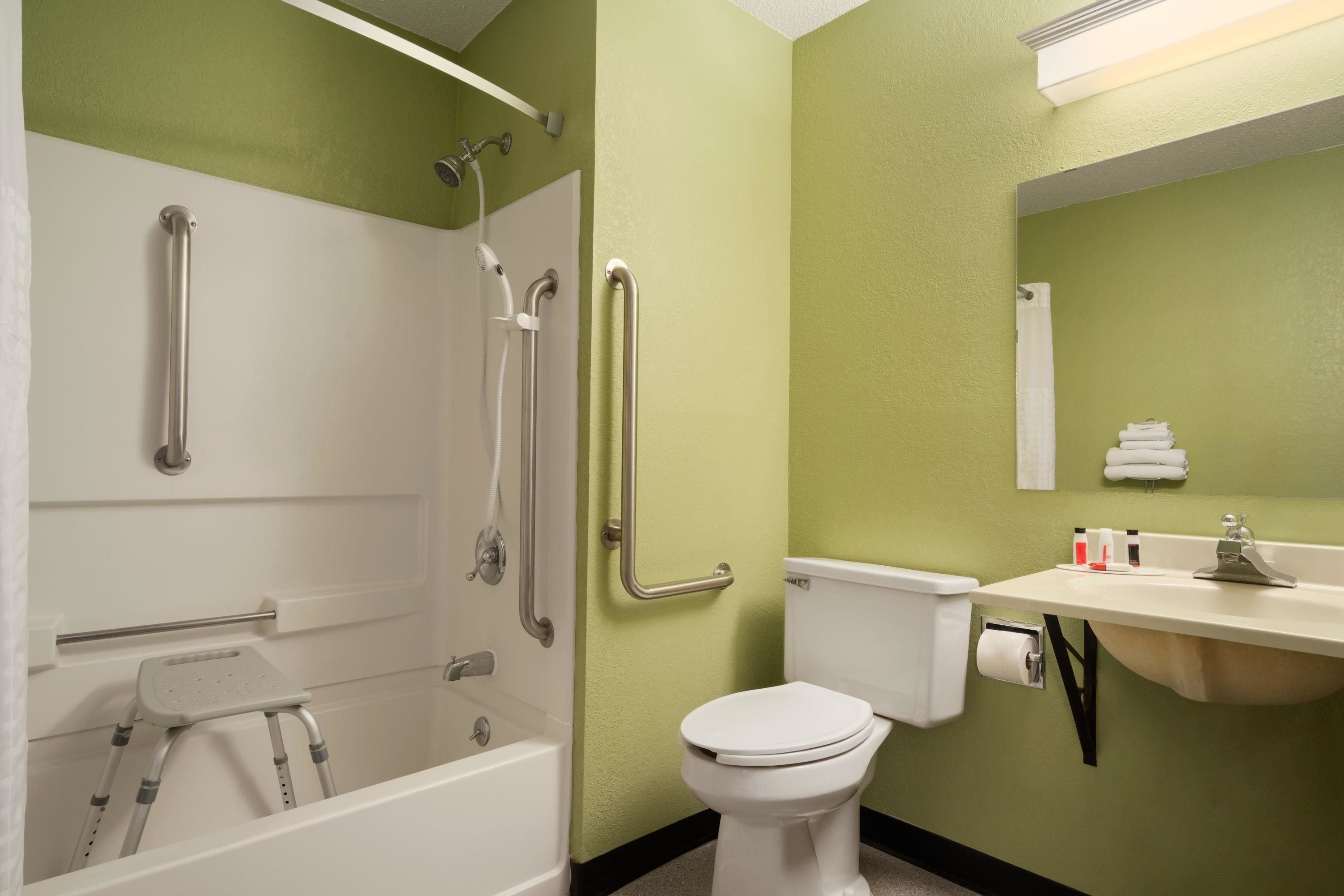 Baño con ducha, inodoro y lavamanos, equipado con barras de apoyo y un taburete.